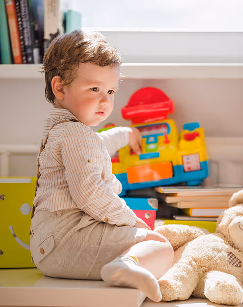 Toddler in beige outfit and yellow socks sitting on a wooden toy with colorful blocks scattered around.